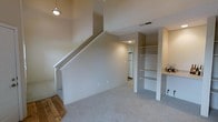 This two-story space offers a practical and open layout designed to maximize functionality. The main level features neutral-toned carpeting that extends throughout the living area, creating a clean and cohesive aesthetic. A staircase with a simple white-painted surround connects the two floors, with a pendant light fixture overhead adding a touch of character to the vertical space.

Built-in open shelving units are prominently featured, providing ample storage and display opportunities without the need for additional furniture. The shelves are finished in white, complementing the light, neutral wall tones throughout. Adjacent to the shelving, a countertop workspace or utility surface offers additional functionality.

A glimpse into the background reveals a corridor leading toward what appears to be a kitchen or utility area, where a white refrigerator is visible, suggesting a well-equipped back section of the space. Recessed lighting illuminates the hallway, while a visible HVAC vent in the ceiling indicates a climate-controlled environment. Overall, the space presents a clean, move-in-ready interior with a straightforward design that emphasizes storage and usability across both levels.