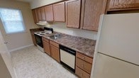 This single-floor residential unit offers a practical and well-appointed living space designed for comfortable everyday use. The kitchen serves as a central highlight, showcasing warm oak cabinetry that extends from floor to ceiling, providing abundant storage for cookware, pantry items, and more. The countertops feature a rich, dark marbled laminate surface that runs the full length of the kitchen, offering generous workspace for meal preparation. Appliances include a white electric range with coil burners, a built-in dishwasher, and a full-size white refrigerator, providing all the essentials for a fully functional kitchen. A stainless steel double-basin sink is conveniently positioned along the counter, complemented by an overhead range hood above the stove. The light-toned tile flooring ties the kitchen together, reflecting light and contributing to a clean, open feel. The neutral wall tones and classic cabinetry create a warm, inviting ambiance throughout. The overall layout is efficient and thoughtfully organized, making the most of the available space while maintaining a tidy and accessible environment suited for a variety of living needs.