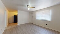 This single-story unit features a straightforward, functional layout distributed across several rooms. The main living area is finished with light gray vinyl plank flooring and neutral white walls, creating a clean and bright atmosphere. A ceiling fan with a built-in light fixture is centrally mounted, providing both ventilation and ambient lighting. A window fitted with horizontal blinds allows natural light to filter into the space, and a door provides direct access to the exterior.

The kitchen is positioned adjacent to the living area and is equipped with warm-toned wood cabinetry on both the upper and lower sections. Black appliances, including a range, dishwasher, and over-the-range microwave, are included. A double-basin stainless steel sink sits beneath a window, adding natural light to the workspace.

The bedrooms are carpeted in a neutral gray tone and each includes a ceiling fan. One bedroom features two closets with sliding doors, offering generous storage, along with a window overlooking greenery outside.

The bathroom includes a single-sink vanity with wood cabinetry, a large mirror, and a full tub and shower combination. Light vinyl plank flooring carries through here as well, maintaining consistency throughout the unit.