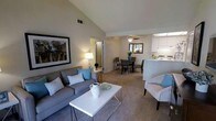 This single-floor space showcases an open-concept floor plan that seamlessly connects the living, dining, and kitchen areas. The living room is anchored by vaulted ceilings that lend a sense of spaciousness and airiness to the space. Soft, neutral-toned carpeting runs throughout, complementing the warm, understated color palette of the walls. The living area comfortably accommodates a full sofa set paired with a clean-lined white coffee table, creating an inviting and functional gathering space. A large framed artwork on the wall adds a decorative focal point.

Flowing naturally from the living room, the dining area features a modest dining table and chairs positioned beneath a ceiling fan, which provides both practical ventilation and ambient lighting. A mirror on the dining area wall enhances the sense of depth and light. Adjacent to the dining space, the kitchen is accessible via a raised bar-style pass-through counter, providing convenience and connectivity. The kitchen features white cabinetry and overhead lighting that brightens the cooking area. A doorway visible from the living room suggests access to a private hallway or additional rooms beyond.