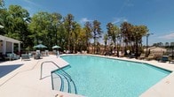 This amenity space combines an expansive outdoor swimming pool with a well-appointed fitness center, providing a well-rounded recreational destination for residents. The outdoor pool area features crystal-clear water in a generously sized pool, surrounded by a wide concrete deck lined with lounge chairs and shaded umbrella seating. A secure perimeter fence encloses the space, while towering pine trees frame the backdrop, creating a private, resort-like atmosphere beneath open blue skies.

The fitness center is accessed through a glass-paneled entry door that overlooks the pool area, creating a seamless connection between indoor and outdoor amenities. Inside, the workout space is bright and modern, featuring a vibrant multi-colored tile floor in shades of teal, green, and gray. The room is equipped with a comprehensive selection of cardio machines, including multiple treadmills and ellipticals, along with strength training equipment and a dedicated free weights area. Large windows along one wall allow for abundant natural light and garden views, while ceiling fans and recessed lighting maintain a comfortable environment. Wall-mounted mirrors and a flat-screen television complete the space, enhancing both functionality and atmosphere for an effective workout experience.