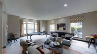 This gameroom offers a well-appointed recreational space designed for both entertainment and relaxation. The room is finished with neutral taupe walls and smooth ceilings with recessed lighting, creating a bright and inviting atmosphere. Multiple windows with patterned drapery and Roman shades line the walls, allowing abundant natural light to filter in while providing views of the surrounding greenery.

The space is anchored by a sleek pool table positioned near the windows, complemented by a shuffleboard table along the opposite wall — both ideal for social gatherings. A central lounge area features a set of contemporary upholstered armchairs in soft gray tones with rust-orange accent pillows, arranged around a round dark-wood coffee table styled with decorative objects. A patterned area rug with warm orange accents grounds the seating arrangement and adds visual interest to the neutral flooring.

A bold abstract painting serves as a focal point on the main wall, adding a touch of artistic character to the space. Overall, the gameroom balances a modern aesthetic with functional recreational amenities, making it a versatile and well-designed communal area.