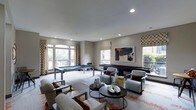 This gameroom offers a well-appointed recreational space designed for both entertainment and relaxation. The room is finished with neutral taupe walls and smooth ceilings with recessed lighting, creating a bright and inviting atmosphere. Multiple windows with patterned drapery and Roman shades line the walls, allowing abundant natural light to filter in while providing views of the surrounding greenery.

The space is anchored by a sleek pool table positioned near the windows, complemented by a shuffleboard table along the opposite wall — both ideal for social gatherings. A central lounge area features a set of contemporary upholstered armchairs in soft gray tones with rust-orange accent pillows, arranged around a round dark-wood coffee table styled with decorative objects. A patterned area rug with warm orange accents grounds the seating arrangement and adds visual interest to the neutral flooring.

A bold abstract painting serves as a focal point on the main wall, adding a touch of artistic character to the space. Overall, the gameroom balances a modern aesthetic with functional recreational amenities, making it a versatile and well-designed communal area.