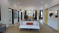 The Tracery is a thoughtfully designed single-story space that blends contemporary elegance with inviting warmth. The main living area immediately captures attention with its open-concept layout, anchored by crisp white walls, crown molding, and recessed lighting that complement the decorative chandelier overhead. Large, floor-to-ceiling windows dressed in rich, dark curtains allow generous natural light to pour in, creating a bright and airy atmosphere throughout.

The space is curated with an eclectic mix of artistic furnishings, including a sleek white settee accented with a rust-colored bolster pillow, sculptural décor pieces, and bold wall art that add character and visual interest. A lounge area in the background features additional seating, reinforcing the space's social and communal nature.

Distinct zones are defined by thoughtful design choices — a rustic sliding barn door in a warm yellow finish provides a charming contrast to the otherwise modern aesthetic, while black-framed interior doors and entryways lend a clean, structured look. The flooring anchors the space with a neutral, textured finish that ties the various areas together seamlessly. Overall, The Tracery offers a refined yet welcoming environment suited for gathering, relaxing, and connecting.