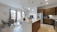 This well-appointed single-floor unit offers a thoughtfully designed open-concept layout that seamlessly connects the kitchen, dining, and living areas. The kitchen is a standout feature, showcasing rich warm-toned cabinetry that extends floor-to-ceiling, a textured herringbone tile backsplash, stainless steel appliances including a French door refrigerator and range, and a generous granite-topped island with bar seating beneath pendant lighting. Light wood-tone flooring runs throughout the space, complementing the bright white walls and creating a cohesive, contemporary aesthetic.

The living area benefits from large floor-to-ceiling windows that flood the space with natural light and offer views of the surrounding neighborhood. A ceiling fan adds functionality to the open living zone. The bedroom is a calm, neutral retreat with clean white walls, light flooring, a clerestory window, and sufficient space for standard furnishings. A short hallway connects the bedroom to the full bathroom, which features warm wood-tone cabinetry, a vanity with storage, and light flooring consistent with the rest of the unit. The overall ambiance is modern, clean, and efficiently designed.