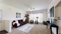 This single-floor residence offers a thoughtfully designed layout that balances comfort and functionality throughout. The generous living room is a standout feature, boasting plush carpeting, crown molding, a ceiling fan with light fixture, and multiple large windows dressed with sheer curtains and blinds that invite an abundance of natural light. A patio door provides direct outdoor access, enhancing the open, airy feel of the space. An elegant arched pass-through connects the living area to the kitchen, which features white cabinetry, dark countertops, rich wood-look flooring, and a full suite of white appliances including a refrigerator with ice maker, electric range, microwave, and dishwasher. A dedicated dining area with crown molding and carpet offers an intimate setting for meals. The bedroom provides a quiet retreat with soft carpet and ample room for furnishings, including workspace. The bathroom impresses with a long double-vanity, white cabinetry, a wide mirror, and wood-look tile flooring, complemented by a separate shower-tub combination. A dedicated laundry room houses a full-size washer and dryer, adding everyday convenience. Neutral tones and crown molding throughout lend the space a clean, cohesive aesthetic.