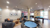 This single-floor entertainment space is thoughtfully designed to accommodate a variety of social and recreational activities. The open-concept layout allows for easy flow between distinct activity zones, each outfitted with dedicated amenities. Recessed lighting throughout the ceiling keeps the space well-illuminated, complemented by natural light streaming in through large windows at the far end of the room.

A central game table with a blue felt surface and built-in cup holders anchors the foreground, while a full-sized billiards table with a teal felt surface occupies an adjacent zone. Ceiling fans are positioned strategically to maintain comfort across the spacious interior. A lounge area features modular gray seating and cylindrical wood accent stools arranged atop a patterned area rug, creating a relaxed gathering spot. Framed graphic artwork lines the white walls, adding a modern, artistic character to the environment.

An accent wall in deep magenta adds a bold visual contrast near a refreshment station equipped with what appears to be a vending machine and bar-height seating. Structural columns punctuate the space without disrupting its open, welcoming ambiance.