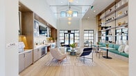 This generously proportioned single-story space is designed as a modern communal lounge and café area, offering a bright, open-plan layout that accommodates a variety of social and relaxation settings. The architecture is immediately distinguished by dramatically soaring ceilings with vaulted detailing, which amplify the sense of volume and airiness throughout the interior.

The entry foyer features wide-plank light wood flooring, crisp white walls, and oversized framed artwork, setting a sophisticated tone. Steel-frame black glass doors and floor-to-ceiling gridded windows are prominent architectural elements throughout, framing outdoor views and flooding the interior with natural light.

The café kitchenette is outfitted with light wood cabinetry, a textured tile backsplash, built-in appliances including a beverage refrigerator, and ample counter space. Built-in open shelving and a mounted television anchor the space functionally.

The living area is organized into distinct zones: a lounge seating area anchored by a statement woven chandelier, a gallery wall with a wall-mounted television, and a cozy window-seat nook set within a teal accent alcove overlooking the outdoors. Herringbone-patterned area rugs define individual seating arrangements, while open shelving units provide display storage. The overall ambiance is contemporary, light-filled, and welcoming.