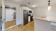 This well-designed 2-bedroom, 1-bathroom unit offers a practical and modern single-floor layout with consistent warm-toned hardwood-style flooring throughout. The open-concept living and dining area is bright and spacious, featuring neutral walls and a door leading to an outdoor patio framed by large windows that invite natural light. The kitchen is efficiently appointed with white shaker-style cabinetry, a peninsula with a built-in sink and pendant lighting overhead, stainless steel refrigerator, electric range, and over-the-range microwave — all complemented by clean countertops and easy access to the main living space. The bathroom is finished with a white vanity, framed mirrors, a glass-enclosed tub and shower combination, and wood-look flooring that ties in with the rest of the unit. Both bedrooms are generously sized with neutral tones and hardwood-style floors. The first bedroom includes a closet, while the second features a large window offering outdoor views and ample natural light. The layout connects all spaces through a central hallway, making the unit feel cohesive and functional throughout.