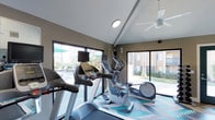 The fitness center at 3675 Barnard in Oceanside is a well-appointed, single-story facility designed to support an active lifestyle. The space features vaulted ceilings that lend an open, airy feel, complemented by a ceiling fan that helps maintain comfortable airflow during workouts. Large windows and sliding glass doors line the walls, flooding the interior with natural light while offering views of the surrounding outdoor area, including what appears to be a pool and landscaped grounds.

The fitness center is equipped with modern cardio machines, including treadmills and elliptical trainers, each outfitted with individual display monitors for entertainment during exercise. The equipment is thoughtfully arranged to maximize usability and movement throughout the space. The flooring features a geometric-patterned design in shades of blue and gray, adding a contemporary visual element to the room.

The neutral-toned walls and clean, minimalist aesthetic create a focused and energizing environment. The combination of natural light, outdoor connectivity, and well-maintained equipment makes this fitness center a functional and inviting space for residents.