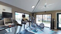 The fitness center at 3675 Barnard in Oceanside is a well-appointed, single-story facility designed to support an active lifestyle. The space features vaulted ceilings that lend an open, airy feel, complemented by a ceiling fan that helps maintain comfortable airflow during workouts. Large windows and sliding glass doors line the walls, flooding the interior with natural light while offering views of the surrounding outdoor area, including what appears to be a pool and landscaped grounds.

The fitness center is equipped with modern cardio machines, including treadmills and elliptical trainers, each outfitted with individual display monitors for entertainment during exercise. The equipment is thoughtfully arranged to maximize usability and movement throughout the space. The flooring features a geometric-patterned design in shades of blue and gray, adding a contemporary visual element to the room.

The neutral-toned walls and clean, minimalist aesthetic create a focused and energizing environment. The combination of natural light, outdoor connectivity, and well-maintained equipment makes this fitness center a functional and inviting space for residents.