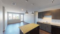 This single-floor residence showcases a thoughtfully designed open-concept layout that seamlessly connects the kitchen and living area. The kitchen serves as a functional centerpiece, featuring a large island topped with speckled granite countertops, dark espresso cabinetry extending to the ceiling, stainless steel appliances including a side-by-side refrigerator with water dispenser, and a built-in microwave. Two dome pendant lights hang above the island, adding a contemporary touch. The adjacent living area features durable wood-look flooring and a sliding glass door leading to a private balcony with exterior views.

The bedrooms are carpeted for comfort, with one bedroom offering direct balcony access through floor-to-ceiling sliding doors and a ceiling fan. Both bedrooms include walk-in closets with built-in rod systems for ample storage. The unit includes multiple bathrooms: one with a tub-shower combination, wood-look tile flooring, and a granite-topped vanity with dark cabinetry; another featuring a single vanity with granite counters and closet access. A compact half-bath with a dark vanity and stained concrete flooring rounds out the space. Neutral gray walls and consistent finishes throughout create a cohesive, modern aesthetic.