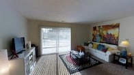 This single-floor space centers around a welcoming living room that draws immediate attention with its large sliding glass door, dressed with vertical blinds that filter natural light throughout the room. The door provides direct access to an outdoor area, offering a seamless connection between interior and exterior. The living room features wall-to-wall carpeting in a neutral tone, which adds warmth underfoot and complements the understated wall color. A sectional sofa anchors the seating area, adorned with an array of colorful accent pillows that introduce playful pops of teal, yellow, and pink. A dark-finished rectangular coffee table sits atop a bold patterned area rug, providing both a functional surface and a striking visual focal point. A piece of vibrant, large-scale artwork on the far wall adds personality and color to the otherwise neutral palette. Along the opposite wall, a white media console provides additional storage and display space. The room's layout is open and functional, with ample space for movement and furnishing. The smooth, light-colored ceiling contributes to an airy atmosphere throughout the living area.