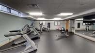 This fitness center is a thoughtfully designed exercise facility that offers a comprehensive range of workout options within a clean, functional space. The room features durable rubber flooring throughout, providing a safe and resilient surface ideal for all types of physical activity. Overhead fluorescent lighting illuminates the entire area evenly, while a row of high clerestory windows along one wall introduces natural light and adds an open feel to the interior.

The cardio section is equipped with Matrix-brand treadmills and elliptical trainers, offering users high-quality equipment for aerobic workouts. A dedicated free weights area includes a well-stocked dumbbell rack with a range of weight options and an adjustable workout bench. Stability balls are available for core training and stretching exercises. Wall-mounted informational charts and workout guides are posted throughout the space, providing helpful references for users.

Large floor-to-ceiling mirrors line multiple walls, visually expanding the room and allowing users to monitor their form during exercise. A wall-mounted flat-screen television adds an entertainment element. A drinking fountain is conveniently located near the exit door. The drop ceiling with textured tiles and neutral grey walls give the facility a clean, professional atmosphere suited for focused, effective workouts.
