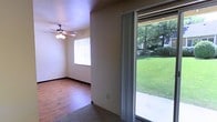 This single-floor space offers a comfortable and functional layout highlighted by an abundance of natural light. The main living area features warm hardwood flooring that transitions to carpet, creating distinct zones within the open space. A ceiling fan with an integrated light fixture is centrally mounted, providing both air circulation and ambient lighting to complement the natural light streaming in through a window fitted with blinds.

A prominent sliding glass door serves as a key architectural feature, offering direct access to the outdoors and framing an unobstructed view of a well-maintained green lawn surrounded by mature trees. The vertical curtains flanking the sliding door allow for flexible light and privacy control. The neutral-toned walls throughout create a clean, versatile backdrop adaptable to various interior styles.

The space benefits from the seamless connection between the interior and the outdoor area, making it feel more expansive than its footprint suggests. The combination of hardwood flooring, natural light, and green outdoor views contributes to a bright, welcoming ambiance suited to everyday living.