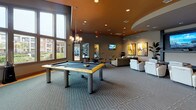 This contemporary clubroom is a well-appointed communal space designed to accommodate a variety of social activities. The open-plan layout is anchored by soaring ceilings with a distinctive warm-toned ceiling detail and recessed lighting, creating an inviting and upscale ambiance throughout.

The main lounge area features plush white leather seating arranged around a central coffee table, complemented by carefully curated abstract artwork on neutral-toned walls. A dedicated screening zone offers rows of premium white leather chairs facing a large wall-mounted flat-screen TV, ideal for group viewings or presentations.

A billiards table takes center stage near floor-to-ceiling windows that flood the space with natural light and provide views of the surrounding exterior. Adjacent dining tables and chairs offer additional seating for gatherings or casual meals.

The fully equipped kitchen is finished with dark espresso cabinetry, granite countertops, and a striking mosaic tile backsplash. Stainless steel appliances, including a dishwasher, under-counter refrigerator, and a coffee station, make this kitchen both functional and visually sophisticated. Tile flooring throughout the kitchen provides a clean contrast to the carpeted lounge areas. Together, these spaces form a cohesive, amenity-rich environment built for community engagement.