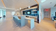 This sophisticated single-floor space is designed with a contemporary open-plan layout that seamlessly blends functionality with modern aesthetics. Light hardwood flooring flows throughout, complementing the bright, white interior and creating a cohesive, airy atmosphere.

Floor-to-ceiling windows dominate the perimeter, flooding the space with abundant natural light and framing sweeping panoramic views of the surrounding cityscape and lush treetops beyond. The elevated vantage point adds a distinctive sense of openness and grandeur.

A central kitchenette and bar area serves as the social hub of the space, featuring a long white countertop with bar-height seating, pendant lighting, dual mounted flat-screen televisions, and a vibrant blue-tiled backsplash that adds a bold design accent. Open shelving and stainless steel appliances complete this well-appointed area.

A casual lounge nook offers a relaxed retreat, furnished with contemporary high-back chairs, a plush area rug, a wall-mounted television, and a slatted wood ceiling detail that adds warmth and texture. Nearby, a bright meeting or dining area features round glass-top tables paired with mixed modern chairs, ideal for collaborative gatherings.

A quieter seating corner with sculptural leather lounge chairs and a wire side table provides an intimate setting for conversation or reflection, all bathed in natural light from expansive windows. The overall ambiance is polished, welcoming, and thoughtfully designed for both work and leisure.