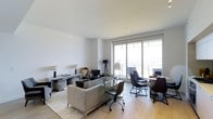 This thoughtfully designed single-floor space presents a seamless open-plan layout that combines living and dining areas into one cohesive, light-filled environment. The centerpiece of the main living area is a large sliding glass door and floor-to-ceiling window system that floods the space with natural light while offering direct access to a private balcony with expansive views beyond. The light hardwood flooring runs throughout, complementing the clean white walls and creating a warm, neutral palette.

The living area is anchored by a plush grey sectional sofa paired with a sleek glass coffee table, accented by stylish floor and table lamps that add layers of ambient lighting. Adjacent to it, the dining zone features a minimalist table surrounded by modern upholstered chairs, perfectly positioned to take advantage of the natural light and outdoor views. The overall aesthetic is contemporary and refined, with a carefully curated mix of textures — from soft furnishings and fur-accented throw pillows to glass and metal accents — that lend the space a sophisticated urban character. Clean lines, an uncluttered layout, and an abundance of natural light define the ambiance, making this a functional yet elegant space.