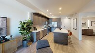 Located at 1919 Pacific Hwy in San Diego, this model unit showcases a thoughtfully designed single-floor layout with contemporary finishes throughout. The open-concept kitchen is a standout feature, outfitted with floor-to-ceiling dark wood cabinetry, white quartz countertops, stainless steel appliances including a gas range, microwave, dishwasher, and refrigerator, and a large kitchen island with bar seating — ideal for both cooking and entertaining. Light wood-style flooring runs throughout the unit, lending warmth and continuity to each space. The living area offers a relaxed, modern atmosphere complemented by curated wall art and recessed lighting. The bedroom is well-appointed with a bright window offering outdoor views, a dedicated walk-in closet with built-in rod and shelving, and ample room for furnishings. The full bathroom features a wood-toned tile surround tub-shower combination, open corner shelving, a backlit vanity mirror, and a sleek floating vanity with dark cabinetry and a white countertop. Clean lines, neutral tones, and modern fixtures define the overall aesthetic, creating a cohesive and sophisticated living environment throughout.
