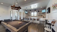 This contemporary amenity space is thoughtfully designed to accommodate a variety of social and recreational activities across a single, open floor plan. The centerpiece of the main gathering area is a full-size pool table positioned beneath a striking industrial-style chandelier crafted from reclaimed wood and metal, anchoring the space with rustic character. Dark hardwood flooring flows throughout, complementing the warm tones of the exposed brick accent wall that lines the wet bar area.

The wet bar is a standout feature, offering an expansive countertop with ample bar seating, stainless steel appliances including built-in microwaves and beverage coolers, open wooden shelving, and multiple wall-mounted televisions. Sculptural pendant lights with a starburst design hang overhead, adding a bold modern touch.

An adjacent lounge area provides plush dark leather seating arranged around natural wood stump side tables and a wall-mounted display screen, creating a relaxed setting for gathering. Nearby, a dining zone features concrete-topped tables with mixed-material chairs set against large windows and glass doors that open to an outdoor terrace, filling the space with natural light. A smaller nook with a bistro table and colorful artwork offers an intimate spot for casual conversation or focused work.