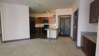 This single-floor residence offers an open-concept layout that seamlessly connects the kitchen and living areas. The kitchen is well-appointed with rich, dark wood cabinetry, black appliances including a refrigerator, microwave, and range, and a tile backsplash that adds texture to the space. A peninsula with a light-toned countertop and bar-height seating serves as a natural divider between the kitchen and the rest of the living area, providing a functional space for casual dining or meal prep. The kitchen flooring features dark tile, which transitions into plush carpeting in the open living area, creating a clear and comfortable separation between the two zones. Track lighting mounted on the ceiling provides focused illumination throughout the kitchen. The entryway, visible from the main living space, features a bold dark-paneled door and tile flooring, lending a clean and defined sense of arrival. Neutral-toned walls throughout keep the space feeling bright and versatile. The thoughtful layout and cohesive design elements make this a practical and inviting living environment.