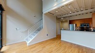 This two-story loft-style residence showcases a thoughtfully designed open-concept layout that blends modern industrial aesthetics with functional living. The main floor features warm hardwood flooring throughout and soaring double-height ceilings with exposed structural beams and track lighting, lending the space an urban, contemporary character. Floor-to-ceiling glass sliding doors flood the living area with natural light and open to a private balcony, offering picturesque views of vibrant greenery and autumn foliage. The well-appointed kitchen is equipped with stainless steel appliances — including a gas range, built-in microwave, and refrigerator — complemented by rich wood cabinetry, dark granite countertops, a tile backsplash, and a peninsula that serves as a natural divider between the kitchen and living area. A full bathroom on the main level features tile flooring, a wood-accented vanity, and a glass-enclosed walk-in shower with a bold red tile accent. Ascending to the second floor, the upper level offers carpeted bedroom space with a walk-in closet providing ample storage, a ceiling fan, and an open railing overlooking the main floor below. A second bathroom with a vanity and access panel completes the upper level, creating a versatile and well-organized two-floor living experience.
