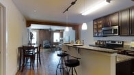This single-floor space exudes rustic charm and warmth through a thoughtful combination of historic architectural elements and contemporary updates. Wide-plank hardwood floors run throughout, lending a sense of continuity and character from room to room. Exposed wooden beams frame the doorways and ceilings, adding structural interest and a nod to the space's original craftsmanship.

The entryway greets visitors with a classic console table and decorative accents, setting a welcoming tone. Moving deeper into the space, the open layout connects a dining area to a comfortable living room, where exposed brick walls create a cozy, textured backdrop. A ceiling fan overhead and natural light filtering through windows enhance the inviting ambiance of the living area.

The kitchen is well-appointed with dark cabinetry, granite countertops, and stainless steel appliances. A breakfast bar with pendant lighting and bar seating serves as a natural transition between the kitchen and the adjoining living and dining spaces, making it ideal for casual meals or entertaining. Track lighting overhead ensures the kitchen remains bright and functional.

Overall, the space masterfully balances the warmth of its original architectural features — exposed brick, reclaimed wood floors, and heavy timber beams — with modern finishes, creating a distinctive and welcoming environment.
