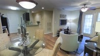This single-floor residence offers a thoughtfully designed open-concept layout that seamlessly connects the main living areas. Warm hardwood flooring runs throughout, lending a cohesive and inviting feel to the entire space.

The dining area is anchored by a round glass-top table surrounded by elegantly upholstered chairs, accented by decorative candle holders that add a refined touch. Adjacent to the dining space, the living room features a comfortable curved accent chair and additional seating, oriented around a wall-mounted flat-screen television set atop a sleek media console. A tall floor lamp provides warm ambient lighting, complementing the recessed ceiling lights found throughout the home.

The kitchen is visible beyond a granite-topped breakfast bar, offering a glimpse of the cabinetry and fixtures within. The raised bar creates a natural boundary between the kitchen and the open living and dining areas while maintaining an airy, connected feel.

Neutral tones on the walls, along with recessed lighting, create a bright and welcoming atmosphere throughout the home. The layout is both functional and stylish, making the most of the available space while ensuring a comfortable flow between rooms.