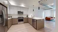 This single-floor residence showcases a cohesive modern design aesthetic defined by a neutral palette, light wood-look flooring, and clean-lined finishes throughout. The kitchen serves as a central highlight, featuring an L-shaped layout with two-tone cabinetry — crisp white upper cabinets paired with warm wood-grain lower cabinets — and light quartz countertops. A peninsula with an integrated sink extends into the living area, topped by sleek black cylindrical pendant lights. Stainless steel appliances, including a French door refrigerator, electric range, over-the-range microwave, and dishwasher, complete the well-equipped space. An exposed metallic duct overhead adds an industrial design accent.

The bedroom is bright and minimalist, featuring large windows with roller shades that allow generous natural light to filter in, contributing to the unit's airy atmosphere. Two bathrooms serve the residence: one featuring a glass-enclosed walk-in shower with vertical subway tile and a wood-toned vanity, and a second bathroom with a matching vanity and clean white walls. Throughout the space, consistent light wood flooring, white walls, and gold-toned hardware details unify the overall contemporary design.