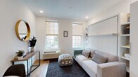 Located at 95 Main St in Hartford, Connecticut, this thoughtfully designed single-floor studio unit maximizes functionality through smart space planning and modern finishes throughout. The open-plan layout seamlessly integrates the living, sleeping, and dining areas, creating a cohesive and versatile environment.

The main living area is anchored by a built-in Murphy bed system with integrated shelving, allowing the space to transition effortlessly between a comfortable lounge and a sleeping area. A light gray sofa, dark area rug, and patterned ottoman complement the neutral white walls and warm-toned luxury vinyl plank flooring. Recessed lighting and two large windows fitted with roller shades bring in natural light while maintaining privacy.

The kitchen features gray shaker-style cabinetry, a white subway tile backsplash, white countertops, and stainless steel appliances including a full-size refrigerator, range, and over-the-range microwave. A compact dining area with a round marble-top table sits adjacent to the kitchen, making efficient use of the available space.

The bathroom continues the clean, modern aesthetic with wood-look flooring, gray vanity cabinetry, a white countertop, and a walk-in shower enclosed by a black-framed glass door. The overall ambiance is bright, minimal, and contemporary.
