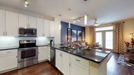 This single-floor residence showcases a thoughtfully designed open-concept layout that seamlessly connects the kitchen and living areas, creating a cohesive and inviting atmosphere. The kitchen is a standout feature, outfitted with crisp white cabinetry, a classic white subway tile backsplash, and dark granite countertops that provide a striking contrast. A central island with an undermount sink doubles as a breakfast bar, ideal for casual dining or entertaining. Stainless steel appliances, including a range, microwave, and dishwasher, complete the modern culinary space. Warm hardwood flooring flows throughout, adding richness and continuity to the interior. Pendant lighting and a track lighting system illuminate the kitchen with a contemporary flair, while a ceiling fan in the living area ensures year-round comfort. The living space is anchored by a bold navy accent wall that adds depth and character, complemented by patterned drapes and a window that draws in natural light. The overall ambiance is modern yet warm, balancing stylish design choices with a comfortable, livable aesthetic that makes this space both functional and visually appealing.