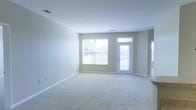 This single-floor space offers a clean, welcoming atmosphere defined by its light-filled and open layout. The main living area is generously proportioned, featuring soft wall-to-wall carpeting in a neutral tone that complements the crisp white ceiling and light gray-beige walls. The space is bathed in natural light, thanks to a large double window fitted with horizontal blinds and a full-length glass-panel exterior door, both of which face outward to what appears to be an outdoor deck or patio area. The glass door allows for seamless visual connection to the outdoors while also serving as an additional source of natural illumination. Subtle architectural details such as clean white baseboards and ceiling-mounted ventilation registers contribute to the space's tidy and well-maintained character. The open floor plan provides significant flexibility for a variety of furniture arrangements and uses. The overall ambiance is calm, airy, and move-in ready, with a neutral color palette that serves as an excellent canvas for personalization. The single-story layout ensures ease of accessibility and flow throughout the space.