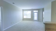 This single-floor space offers a clean, welcoming atmosphere defined by its light-filled and open layout. The main living area is generously proportioned, featuring soft wall-to-wall carpeting in a neutral tone that complements the crisp white ceiling and light gray-beige walls. The space is bathed in natural light, thanks to a large double window fitted with horizontal blinds and a full-length glass-panel exterior door, both of which face outward to what appears to be an outdoor deck or patio area. The glass door allows for seamless visual connection to the outdoors while also serving as an additional source of natural illumination. Subtle architectural details such as clean white baseboards and ceiling-mounted ventilation registers contribute to the space's tidy and well-maintained character. The open floor plan provides significant flexibility for a variety of furniture arrangements and uses. The overall ambiance is calm, airy, and move-in ready, with a neutral color palette that serves as an excellent canvas for personalization. The single-story layout ensures ease of accessibility and flow throughout the space.