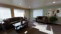 This single-story space showcases a well-appointed living room as its centerpiece. The room is finished with wall-to-wall carpet in a warm brown tone, providing a comfortable and cohesive foundation throughout. Two generously sized windows dressed with white horizontal blinds allow abundant natural light to filter into the space, casting bright patterns across the floor and creating an inviting, airy atmosphere. Recessed lighting in the textured ceiling supplements the natural light, ensuring the room remains well-lit at all hours.

The walls are painted in a neutral tan and soft sage green combination, lending a calm, understated character to the interior. A standard white paneled door is centrally positioned between the two windows, offering a clean architectural focal point. The living area comfortably accommodates two dark brown upholstered sofas arranged around a rectangular dark wood coffee table, highlighting the room's practical and spacious layout. A small accent table with a lamp adds a functional decorative touch near the entry door. Overall, the space presents a clean, functional, and comfortable living environment with a relaxed, approachable aesthetic.