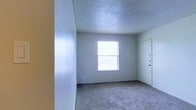 This single-story space presents a straightforward and functional interior, as seen in the main room captured in the image. The room features wall-to-wall carpeting in a neutral tone, providing a comfortable underfoot surface that also contributes to a quiet atmosphere. The walls are finished with a textured surface, adding subtle visual depth, while the ceiling carries a popcorn texture treatment that is consistent with the overall utilitarian aesthetic of the space.

A single window positioned along the far wall allows natural light to filter into the room, brightening the interior and creating an open feel despite the modest scale. The white-painted door visible in the corner appears to be a closet or secondary access point, offering practical storage or passage options. The walls are painted in a light, neutral gray-white tone, keeping the space feeling clean and versatile for various uses.

The overall ambiance is understated and move-in ready, with a simple palette that provides a blank canvas for personalization. The single-floor layout ensures easy accessibility throughout, making it a practical and straightforward living or working environment.