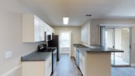 This single-floor residence offers a thoughtfully designed layout centered around a galley-style kitchen that serves as the heart of the home. The kitchen is outfitted with sleek black stainless steel appliances, including a refrigerator, electric range with microwave above, and a dishwasher, providing a full suite of modern conveniences. White shaker-style cabinetry lines both sides of the kitchen, offering generous storage space, while dark granite-look countertops provide a striking contrast. A double-basin sink is positioned along the right counter run, complemented by a brushed nickel faucet. The flooring throughout features light wood-look planks that add warmth and visual continuity to the space. A textured ceiling with recessed lighting and a central skylight panel ensures the kitchen remains bright and well-lit. Natural light filters in through a window at the end of the galley and additional windows visible in adjacent areas, contributing to an open and airy atmosphere. The neutral wall tones in soft beige create a clean, versatile backdrop that complements the overall design. A peninsula counter extension at one end offers additional workspace or casual dining potential, rounding out this functional and attractive kitchen space.