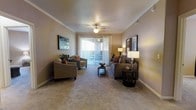 This single-story space features a well-appointed living room that serves as the focal point of the layout. The room is finished with neutral-toned carpeting and warm beige walls that create a welcoming, cohesive atmosphere throughout. Crown molding lines the ceiling, adding a refined architectural detail that elevates the overall aesthetic.

A ceiling fan with a built-in light fixture is centrally positioned, providing both functional ventilation and ambient lighting. Natural light fills the space through a large sliding glass door fitted with vertical blinds, offering access to an outdoor area and bringing in a bright, airy quality. The door serves as a natural focal point along the far wall, drawing the eye and enhancing the sense of openness.

The living area is generously proportioned, comfortably accommodating a full seating arrangement including a sofa, armchairs, and a central coffee table. Framed artwork on opposing walls adds visual balance. Recessed HVAC vents are integrated into the ceiling, reflecting a practical and thoughtfully designed interior. The overall ambiance is calm, neutral, and move-in ready.
