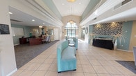 This single-story lobby space makes an immediate impression with its soaring vaulted ceilings and abundant natural light streaming through a dramatic arched window at the entrance. The open layout is both functional and inviting, creating a sense of spaciousness that carries throughout the interior. Large, light-toned floor tiles run across the main area, reflecting natural light and enhancing the bright, clean ambiance.

The seating area is anchored by a curved, teal upholstered bench paired with sleek, modern round accent tables, offering a comfortable and visually cohesive waiting area. The color palette draws from cool tones — soft teals, whites, and warm neutrals — lending the space a calm, professional atmosphere. Recessed lighting and pendant fixtures complement the natural light, ensuring the interior remains well-illuminated throughout the day.

To one side, a warm wood-toned reception desk sits beneath its own dedicated lighting, creating a defined, professional workspace. On the opposite side, a striking mosaic tile accent wall in shades of gray, blue, and brown adds a bold design element, flanked by decorative vases that bring a refined, finishing touch. The overall design balances contemporary style with approachable warmth, making this lobby an excellent first impression for any visitor.