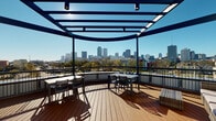 Located on the fourth floor in the heart of New Orleans, LA, this terrace offers an impressive combination of open-air outdoor space and a refined interior lounge. The outdoor deck is finished with warm hardwood decking and follows a graceful curved layout, creating an inviting, sweeping feel. A bold navy-blue steel pergola frames the space above, providing partial shade while allowing abundant natural light to pour in throughout the day. Seating areas with tables and chairs are arranged to take full advantage of the spectacular panoramic views of the New Orleans skyline, with the city's iconic towers and lush tree canopy stretching across the horizon. A secure glass-panel door connects the terrace to the interior, maintaining a seamless transition between indoors and out. The interior lounge area features light hardwood-style flooring, soft neutral gray walls, and warm pendant lighting overhead. Comfortable orange accent chairs arranged around a low coffee table create an inviting seating nook, while framed artwork and a potted plant add understated character. Elevator access is conveniently located within the interior, making this fourth-floor terrace easily accessible and well-suited for relaxed gatherings or enjoying the vibrant New Orleans cityscape.