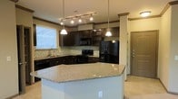 This single-story space centers around a thoughtfully designed kitchen that combines style and functionality. The kitchen is anchored by a large island featuring light-toned granite countertops with subtle flecking, providing generous prep and gathering space. Pendant lights hang elegantly above the island, complementing a track lighting system overhead that ensures thorough illumination throughout the area.

The cabinetry is finished in a rich, dark espresso tone, creating a striking contrast against the lighter countertops and walls. Upper cabinets include glass-front display sections, adding visual interest and an opportunity to showcase decorative items. A mosaic tile backsplash in neutral earth tones runs along the walls, adding texture and detail to the space.

Black appliances, including a French door refrigerator, range, microwave, and dishwasher, contribute to a cohesive, contemporary aesthetic. A window above the sink allows natural light to filter into the kitchen. Crown molding accents the upper cabinetry and structural column, lending an elevated, finished appearance. Light-toned tile flooring runs throughout, tying the design elements together seamlessly.