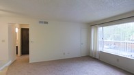 This single-floor space features a spacious, open room finished in a clean, neutral color palette that creates a bright and airy atmosphere. The room is outfitted with wall-to-wall carpet in a light beige tone, adding warmth underfoot and contributing to the overall understated aesthetic. Walls are painted in a soft white, keeping the space feeling open and versatile.

A large window dressed with sheer white curtains allows generous natural light to fill the room, enhancing its sense of openness. A curtain rod is in place, offering flexibility for additional window treatments. The textured popcorn ceiling adds a classic residential character while also housing a centrally positioned HVAC vent, ensuring climate control throughout the space.

An interior door is visible along one wall, suggesting access to an adjoining room or closet, contributing to the functional layout of the space. Electrical outlets are positioned along the baseboards, providing practical utility throughout. The clean white baseboards frame the room neatly, complementing the overall light and minimal design. Overall, the space offers a simple, move-in-ready environment with a calm, neutral ambiance.