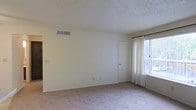 This single-floor space features a spacious, open room finished in a clean, neutral color palette that creates a bright and airy atmosphere. The room is outfitted with wall-to-wall carpet in a light beige tone, adding warmth underfoot and contributing to the overall understated aesthetic. Walls are painted in a soft white, keeping the space feeling open and versatile.

A large window dressed with sheer white curtains allows generous natural light to fill the room, enhancing its sense of openness. A curtain rod is in place, offering flexibility for additional window treatments. The textured popcorn ceiling adds a classic residential character while also housing a centrally positioned HVAC vent, ensuring climate control throughout the space.

An interior door is visible along one wall, suggesting access to an adjoining room or closet, contributing to the functional layout of the space. Electrical outlets are positioned along the baseboards, providing practical utility throughout. The clean white baseboards frame the room neatly, complementing the overall light and minimal design. Overall, the space offers a simple, move-in-ready environment with a calm, neutral ambiance.