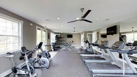 This fitness center offers a comprehensive workout environment designed to accommodate a wide range of exercise routines. The space is laid out on a single floor, with a generous open floor plan that allows for easy movement between equipment stations. Along one side, a row of treadmills provides ample cardio options, complemented by elliptical trainers and stationary bikes positioned throughout the room. A dedicated free weights area features a rack of dumbbells and a flat bench, ideal for strength training. Wall-mounted mirrors along one side enhance the sense of space while allowing users to monitor their form. Two ceiling fans with integrated lighting ensure comfortable air circulation during workouts, and recessed lighting throughout keeps the room well-lit at all times. Large windows along one wall invite natural light and offer views of the surrounding greenery outside. Wall-mounted televisions are positioned for visibility from the cardio equipment, adding to the convenience of longer workout sessions. The flooring consists of durable, cushioned gray carpet suited for a fitness environment. The neutral gray walls and clean, modern finishes contribute to an organized and motivating atmosphere throughout the space.