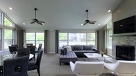 This single-floor space showcases a thoughtfully designed open-concept layout that seamlessly blends a living area with a dining space. Large picture windows line the walls, flooding the interior with generous natural light while offering views of the surrounding greenery and outdoor environment. Gray drapes frame the windows, adding a polished, cohesive aesthetic.

The living area is anchored by a tufted gray sectional sofa paired with a sleek white coffee table, creating a comfortable yet contemporary focal point. A modern white accent chair adds contrast and visual interest. Two stylish ceiling fans with palm-leaf-inspired blades hang overhead, contributing both function and a subtle design element to the space.

Adjacent to the living area, a dining zone features dark chairs arranged around a white table, maintaining the space's neutral, modern color palette. Neutral-toned carpet runs throughout, tying the areas together harmoniously. The overall ambiance is clean, airy, and inviting, with a sophisticated gray-and-white color scheme that lends the space a refined, contemporary character well-suited for both relaxation and social gatherings.