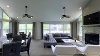 This single-floor space showcases a thoughtfully designed open-concept layout that seamlessly blends a living area with a dining space. Large picture windows line the walls, flooding the interior with generous natural light while offering views of the surrounding greenery and outdoor environment. Gray drapes frame the windows, adding a polished, cohesive aesthetic.

The living area is anchored by a tufted gray sectional sofa paired with a sleek white coffee table, creating a comfortable yet contemporary focal point. A modern white accent chair adds contrast and visual interest. Two stylish ceiling fans with palm-leaf-inspired blades hang overhead, contributing both function and a subtle design element to the space.

Adjacent to the living area, a dining zone features dark chairs arranged around a white table, maintaining the space's neutral, modern color palette. Neutral-toned carpet runs throughout, tying the areas together harmoniously. The overall ambiance is clean, airy, and inviting, with a sophisticated gray-and-white color scheme that lends the space a refined, contemporary character well-suited for both relaxation and social gatherings.