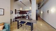 This contemporary loft apartment in downtown Minneapolis offers a perfect blend of industrial charm and modern comfort. The 820 square foot space features an open floor plan with high ceilings and exposed ductwork, creating an expansive feel. Large windows flood the living area with natural light, offering views of the surrounding cityscape.

The living room is a focal point, boasting a vibrant blue rug that adds a pop of color to the neutral palette. A sleek entertainment center and comfortable seating create an inviting atmosphere for relaxation or entertaining. The adjacent kitchen is equipped with stainless steel appliances, dark wood cabinetry, and a central island with bar seating, ideal for casual dining or meal preparation.

The primary bedroom, measuring 150 square feet, features a striking four-poster bed and sliding glass doors, likely leading to a closet or storage area. The second bedroom offers a cozy retreat with stylish furnishings and ample natural light. The bathroom showcases a modern vanity with rich wood tones and a large mirror, complemented by tasteful artwork.

Throughout the loft, the interplay of textures - from smooth concrete floors to exposed brick walls - adds depth and character to the space. Thoughtful decor elements, including potted plants and artwork, soften the industrial edges and create a harmonious living environment. This loft epitomizes urban living, combining functionality with aesthetic appeal in the heart of Minneapolis.