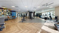 Located at 5121 E La Puente Ave in Phoenix, AZ, this generously sized fitness center offers a well-appointed, single-floor exercise space designed to accommodate a wide variety of workout needs. The room is finished with striking herringbone-patterned hardwood flooring that transitions seamlessly into rubberized mat zones, providing both aesthetic appeal and functional safety for strength training areas.

The facility is equipped with an extensive selection of cardio machines, including treadmills, ellipticals, rowing machines, and stationary bikes. The strength training section features a multi-functional cable machine, a heavy bag, weight benches, a full rack of medicine balls and dumbbells, and additional selectorized strength equipment. Large mirrors line portions of the walls, enhancing the sense of space and supporting proper form during workouts.

Natural light pours in through multiple windows distributed throughout the room, complemented by a drop-panel ceiling with recessed and flush-mounted lighting fixtures, as well as ceiling fans that ensure consistent air circulation. Accent walls in bold blue add a modern, energetic atmosphere to the space. Thoughtful touches such as decorative wall art, indoor plants, and a small lounge seating area contribute to an inviting and motivating environment for users of all fitness levels.