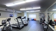 This fitness center offers a comprehensive workout environment designed to accommodate a variety of exercise routines. The space is anchored by multiple treadmills positioned prominently for cardio training, complemented by an elliptical machine and additional cardio equipment. A multi-station strength training machine is situated along one wall, providing users with a range of resistance and weight-based exercises. The open floor area is covered with protective rubber matting, creating a safe and functional surface ideal for stretching, bodyweight exercises, and free-form workouts. Exercise accessories, including stability balls in multiple sizes, are available throughout the space, adding versatility to workout options. The room is illuminated by a series of bright fluorescent overhead light fixtures mounted to a textured ceiling, ensuring consistent and even lighting throughout. Ventilation is supported by ceiling vents, maintaining a comfortable environment during use. A wall-mounted television screen is present, providing entertainment or instructional content for users. A window fitted with vertical blinds allows for natural light to supplement the interior lighting. The neutral-toned walls and dark flooring create a focused, functional atmosphere well-suited for fitness activity.