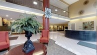 This impressive lobby offers a grand, open atmosphere defined by its soaring double-height ceilings and an open mezzanine level above, framed by ornamental metal railings. A bold, cylindrical column in a rich terracotta hue anchors the center of the space, lending architectural character and visual interest. The flooring transitions between large-format white tile and patterned carpet areas, delineating distinct functional zones throughout the lobby.

A welcoming reception desk is positioned toward the right side of the space, providing a clear point of contact for visitors upon entry. The seating area features a classic upholstered armchair in warm rust tones, complemented by a polished wooden round table and an ornate urn-style planter filled with lush, tropical foliage, adding a touch of natural elegance.

Along the far wall, a gallery-style display of framed artwork is evenly spaced, contributing to the refined and curated aesthetic of the space. Recessed and overhead lighting illuminates the lobby evenly, while a decorative wall clock adds a functional yet stylish accent. The overall ambiance is professional, spacious, and inviting, making it well-suited as a central gathering and reception area for guests and visitors.