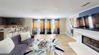 This single-story clubhouse offers a welcoming and functional space designed to serve as a central hub for community members. The main lounge area is generously sized, featuring warm-toned beige walls, light hardwood-style flooring, and recessed lighting that creates a bright and inviting atmosphere. Large windows dressed with bold tri-color curtains in gold, gray, and black allow natural light to flow throughout the space. The lounge is furnished with a charcoal sectional sofa, patterned accent chairs with gold throw pillows, a geometric multi-toned area rug, and a glass coffee table. A fireplace with a white mantel anchors one end of the room, complemented by colorful abstract wall art that adds a contemporary touch. Crown molding details throughout elevate the overall finish of the space. The fully equipped kitchen features dark wood cabinetry, light quartz countertops, stainless steel appliances including a refrigerator with water dispenser, electric range, and built-in microwave, as well as a deep double sink. A small bistro-style table with a chess set provides a casual nook for games and informal gatherings. The clubhouse blends functional amenities with thoughtful design, making it a versatile and comfortable space for residents to socialize and unwind.